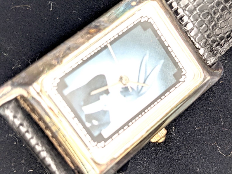 Pu&ograve; includere: Orologio da polso rettangolare argentato con quadrante azzurro e cinturino in pelle nera. L'orologio ha lancette color oro e una corona color oro. Il quadrante ha un design decorativo. Il cinturino ha un motivo strutturato.