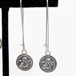 May include: Silver dangle earrings. Each earring has a long, thin wire with a small, round charm. The charm features a detailed image of an angel. The earrings are shown against a white background.