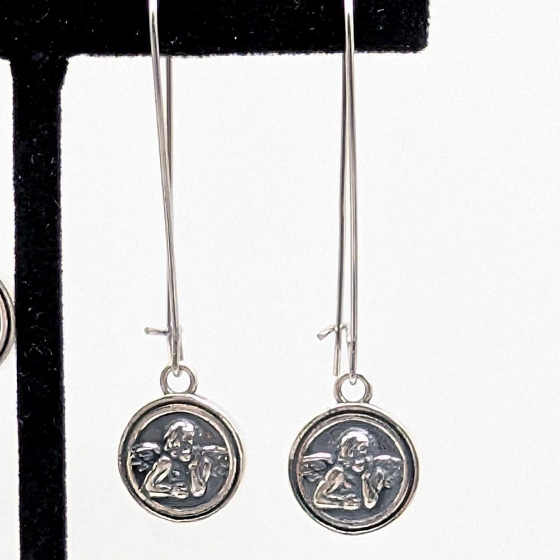 May include: Silver dangle earrings. Each earring has a long, thin wire with a small, round charm. The charm features a detailed image of an angel. The earrings are shown against a white background.