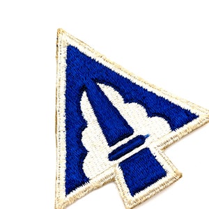 May include: A blue and white embroidered patch with a stylized arrow design. The arrow is pointing to the right and has a white outline. The patch is surrounded by a tan border.