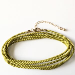 Puede incluir: Un collar de cordón verde oliva de varias hebras con textura de cuerda trenzada. El collar incluye un cierre de mosquetón metálico y una cadena extensora ajustable con una pequeña cuenta redonda al final, en tono dorado pulido.