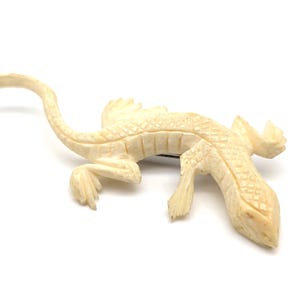 May include: An off-white lizard-shaped brooch featuring a carved, textured scale pattern across its back and head. The piece has a long, curved tail, four distinct legs, and a pin mechanism visible on the underside of the lizard's body.