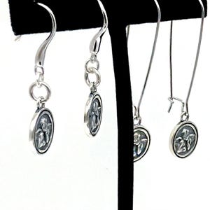 May include: Silver dangle earrings with oval pendants, each displaying a detailed relief design. The earrings are presented on a black stand against a white backdrop, highlighting their intricate design and elegant style.