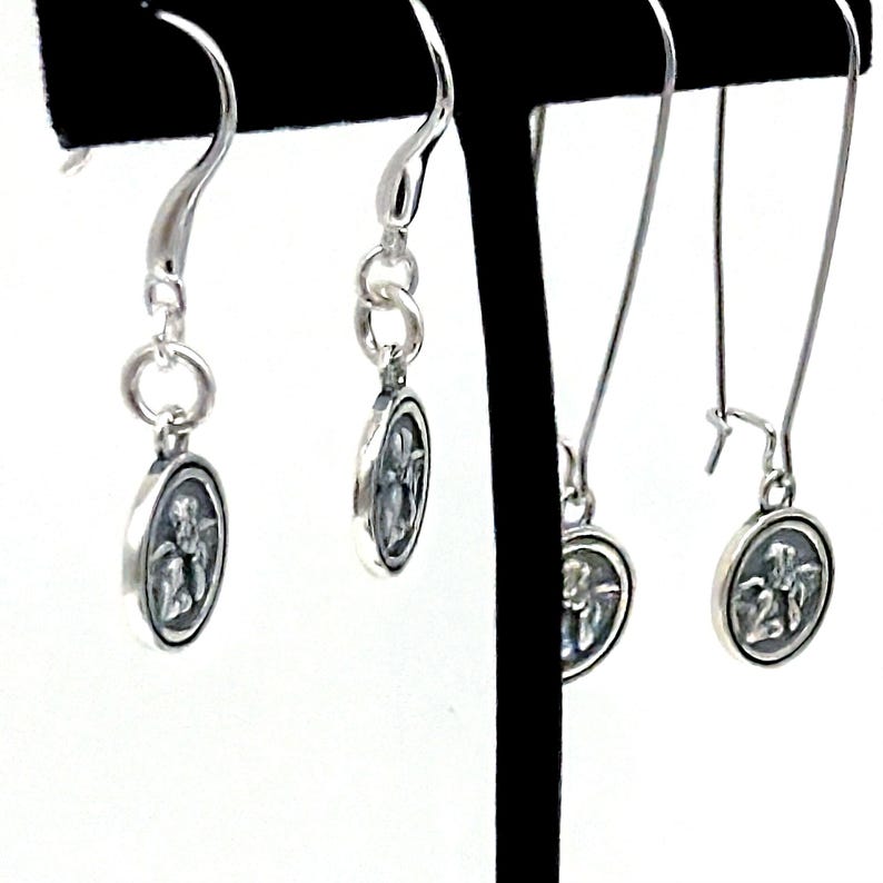 May include: Silver dangle earrings with oval pendants, each displaying a detailed relief design. The earrings are presented on a black stand against a white backdrop, highlighting their intricate design and elegant style.
