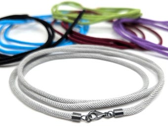 Puede incluir: Un collar de cuerda gris claro con un cierre plateado está en foco, con otras cuerdas coloridas en el fondo. El collar está hecho de un material tejido. Otros colores incluyen negro, azul, verde, morado y burdeos.