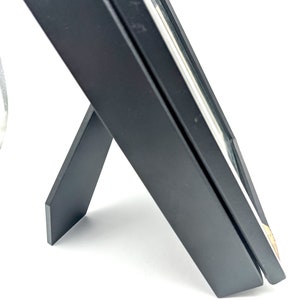 Pu&ograve; includere: Una cornice nera con un cavalletto integrato, angolata su una superficie bianca. La cornice ha un design semplice e moderno con un sottile accento dorato sul bordo. La cornice &egrave; progettata per stare in piedi.