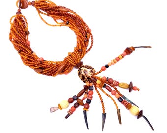 Peut inclure: Un collier artisanal composé de plusieurs rangées de perles orange et marron. Le collier comprend des perles en bois, des éléments en coquillage et des éléments décoratifs. Il est fermé par un fermoir à bascule.