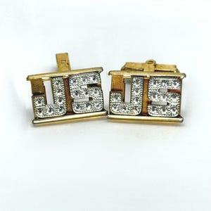 May include: A pair of gold-toned cufflinks featuring the letters JS in a block font. The letters are encrusted with small, clear, sparkling stones set against a metallic gold frame with a horizontal bar design at the top and bottom edges.