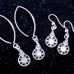 Puede incluir: Pendientes plateados con diseño de rosa de los vientos. Los pendientes tienen una parte superior decorativa y un colgante circular con una rosa de los vientos. Los pendientes están sobre un fondo negro. Son ligeros y cuelgan.