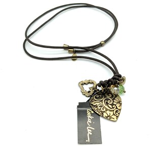 Pode incluir: Um colar de cordão marrom com um pingente de coração dourado e contas verdes. O pingente tem um design floral intrincado. O colar tem uma etiqueta "cookie lee.".