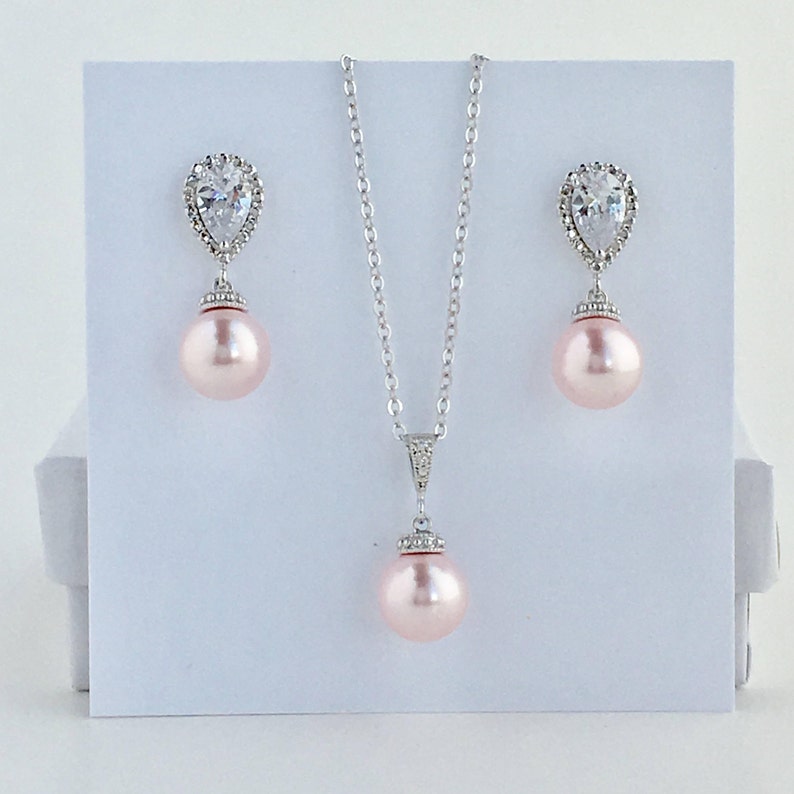 May include: A delicate bridal necklace and earrings set featuring a pear-shaped crystal with a halo of smaller crystals and a pink pearl pendant.