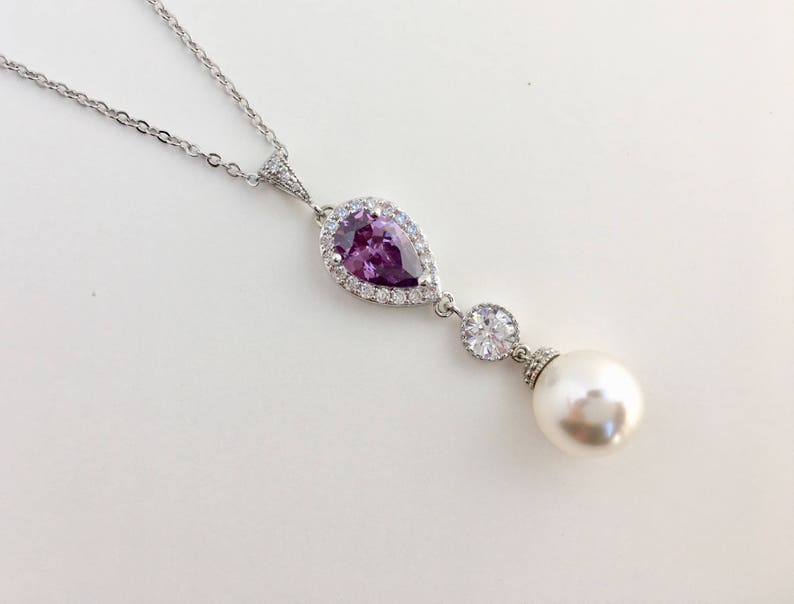 May include: A silver necklace with a pear-shaped purple gemstone surrounded by clear stones and a single pearl pendant.