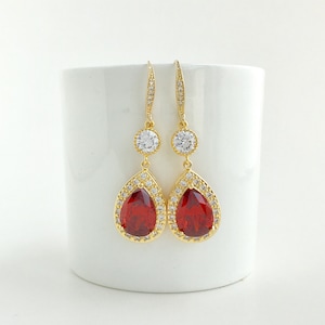 May include: Gold dangle earrings with a teardrop-shaped red gemstone surrounded by smaller clear stones. The earrings have a smaller round clear stone above the red gemstone.