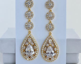 May include: Gold dangle earrings with a pear-shaped crystal surrounded by smaller crystals. The earrings have three round crystal accents above the pear-shaped crystal.