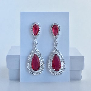 May include: A pair of dangle earrings with a teardrop-shaped red crystal surrounded by a halo of smaller clear crystals.