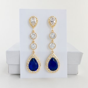 May include: Gold tone dangle earrings with a teardrop shaped blue stone surrounded by smaller clear stones. The earrings have three clear round stones above the blue stone.