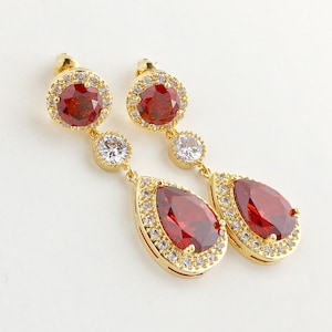 Peut inclure: Boucles d'oreilles pendantes dorées avec une grosse pierre rouge en forme de goutte d'eau entourée d'un halo de petits cristaux transparents. Les boucles d'oreilles ont également une petite pierre rouge et un cristal transparent en haut.