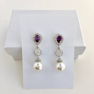 May include: A pair of silver dangle earrings with a pear-shaped purple gemstone, a round clear rhinestone, and a white pearl drop.