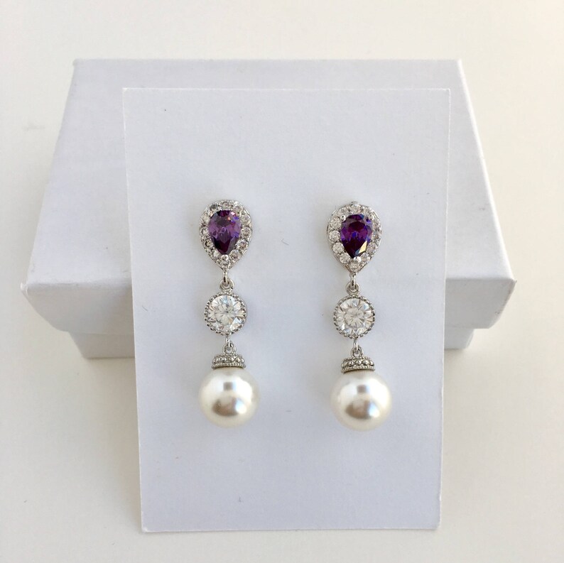 May include: A pair of silver dangle earrings with a pear-shaped purple gemstone, a round clear rhinestone, and a white pearl drop.