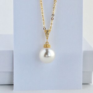 Puede incluir: Un collar de cadena dorada con un colgante de una sola perla blanca grande. La perla est&aacute; engastada en un bisel dorado con peque&ntilde;as piedras transparentes alrededor del borde.
