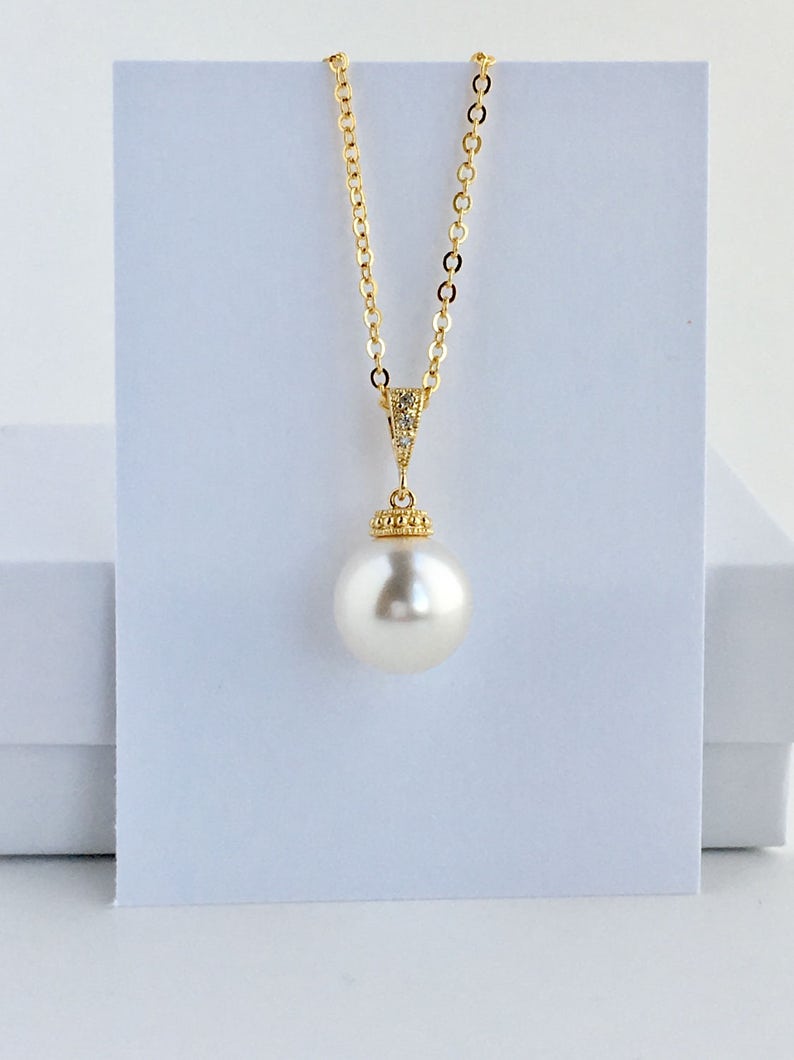 Puede incluir: Un collar de cadena dorada con un colgante de una sola perla blanca grande. La perla est&aacute; engastada en un bisel dorado con peque&ntilde;as piedras transparentes alrededor del borde.