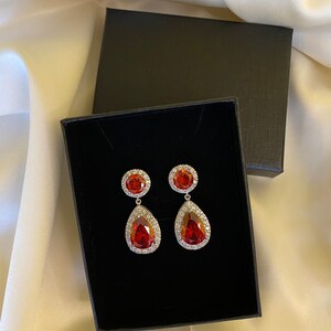 May include: A pair of red teardrop-shaped earrings with a clear crystal halo around each stone. The earrings are in a black jewelry box.