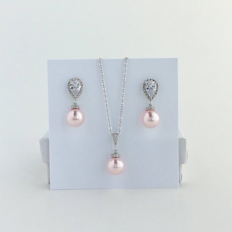 May include: A delicate bridal jewellery set featuring a pear-shaped crystal pendant and matching earrings. The set includes a silver chain necklace and pink pearl accents.