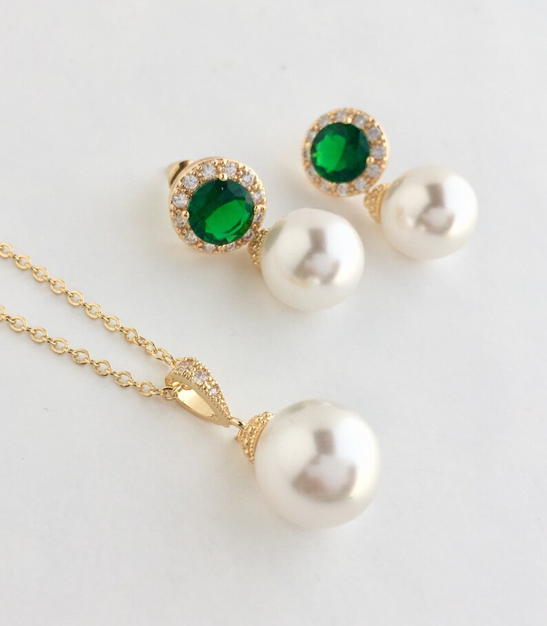 Puede incluir: Un conjunto de collar y pendientes de oro con un gran colgante de perla blanca y pendientes a juego. Los pendientes presentan una piedra preciosa verde rodeada de piedras transparentes m&aacute;s peque&ntilde;as.
