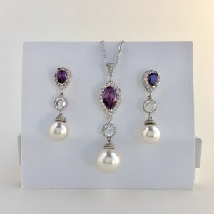May include: A silver necklace and earrings set with a pear-shaped purple gemstone surrounded by smaller clear stones. The necklace and earrings each have a pearl drop.