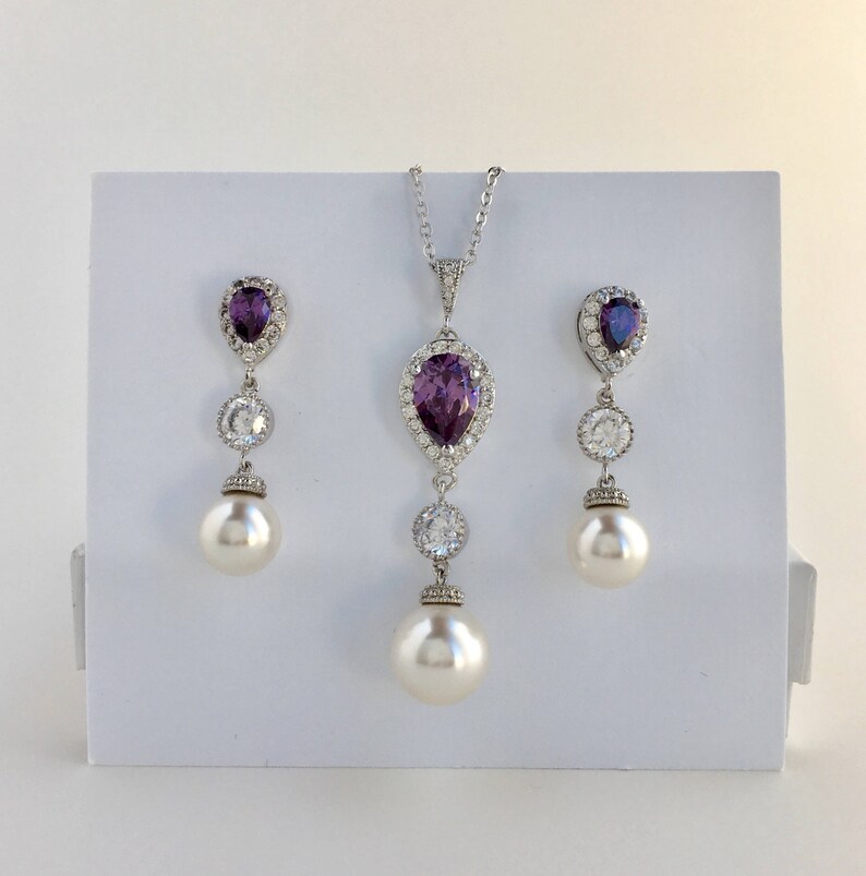 May include: A silver necklace and earrings set with a pear-shaped purple gemstone surrounded by smaller clear stones. The necklace and earrings each have a pearl drop.