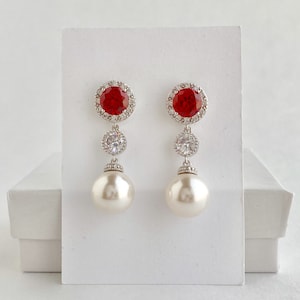 May include: A pair of silver dangle earrings with a large red gemstone surrounded by smaller clear stones, a smaller clear stone, and a large white pearl.