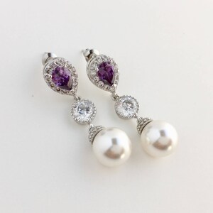 May include: A pair of silver earrings with a pear-shaped purple gemstone, a round clear crystal, and a white pearl drop.