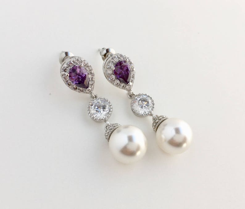 May include: A pair of silver earrings with a pear-shaped purple gemstone, a round clear crystal, and a white pearl drop.