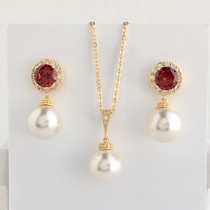 Puede incluir: Un collar y pendientes de oro con una piedra roja rodeada de brillantes transparentes y un gran colgante de perla blanca.