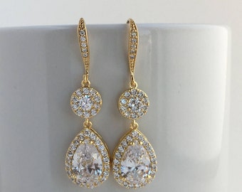 May include: A pair of gold-coloured drop earrings. Each earring has a round cluster of small, clear crystals above a larger, pear-shaped crystal, also surrounded by smaller crystals. The earrings hang from a delicate, crystal-studded hook.
