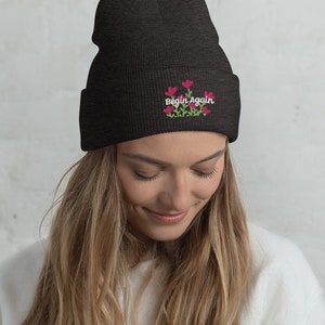 May include: A black beanie with a white embroidered design that says "Begin Again" with pink flowers.
