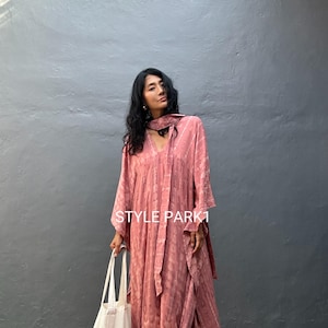 Könnte beinhalten: Ein langes, fließendes, staubrosa Kleid mit V-Ausschnitt und langen Ärmeln. Das Kleid hat ein dezentes Muster und einen passenden Schal. Eine weiße Tragetasche wird in der linken Hand gehalten. Der Text "STYLE PARK1" ist sichtbar.