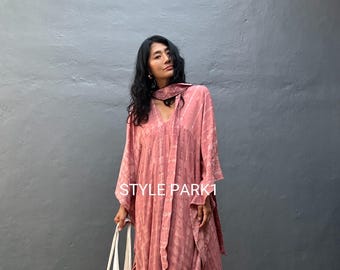 May include: A long, flowing, dusty rose dress with a V-neck and long sleeves. The dress has a subtle pattern and a matching scarf. A white tote bag is held in the left hand. The text "STYLE PARK1" is visible on the dress.