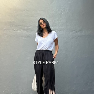 May include: A woman wearing a white v-neck t-shirt and black wide-leg pants with a drawstring waist. The pants have a subtle tie-dye effect. The woman is standing in front of a gray wall. The text "STYLE PARK1" is visible on the image.