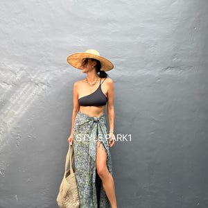 May include: A woman wearing a black one-shoulder swimsuit top and a green and white patterned sarong skirt. She is also wearing a wide-brimmed straw hat and a gold chain necklace. The skirt is tied at the waist and has a slit up the side. She is holding a woven tote bag.
