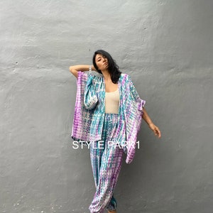 以下が含まれることがあります： 白のタンクトップと金のサンダルを履いた女性が、タイダイ柄のパンツスーツを着ています。パンツスーツはゆったりとしたシルエットで、ワイドレッグスタイルです。画像には「STYLE PARK 1」というテキストが表示されています。