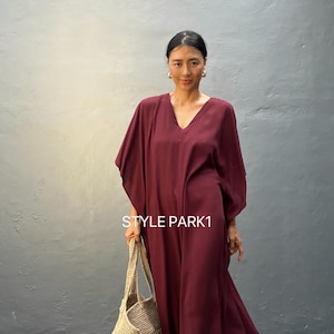 Pode incluir: Uma mulher vestindo um vestido longo de cor bordô com decote em V e caimento solto. Ela está segurando uma bolsa de mão de crochê bege. O texto "STYLE PARK1" é visível no vestido.