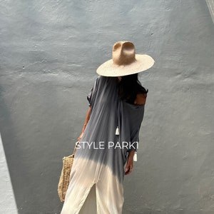 May include: A woman wearing a gray and white ombre maxi dress with a straw hat and a woven tote bag. The dress has a slit up the front and tassels on the neckline. The text "STYLE PARK1" is visible on the dress.