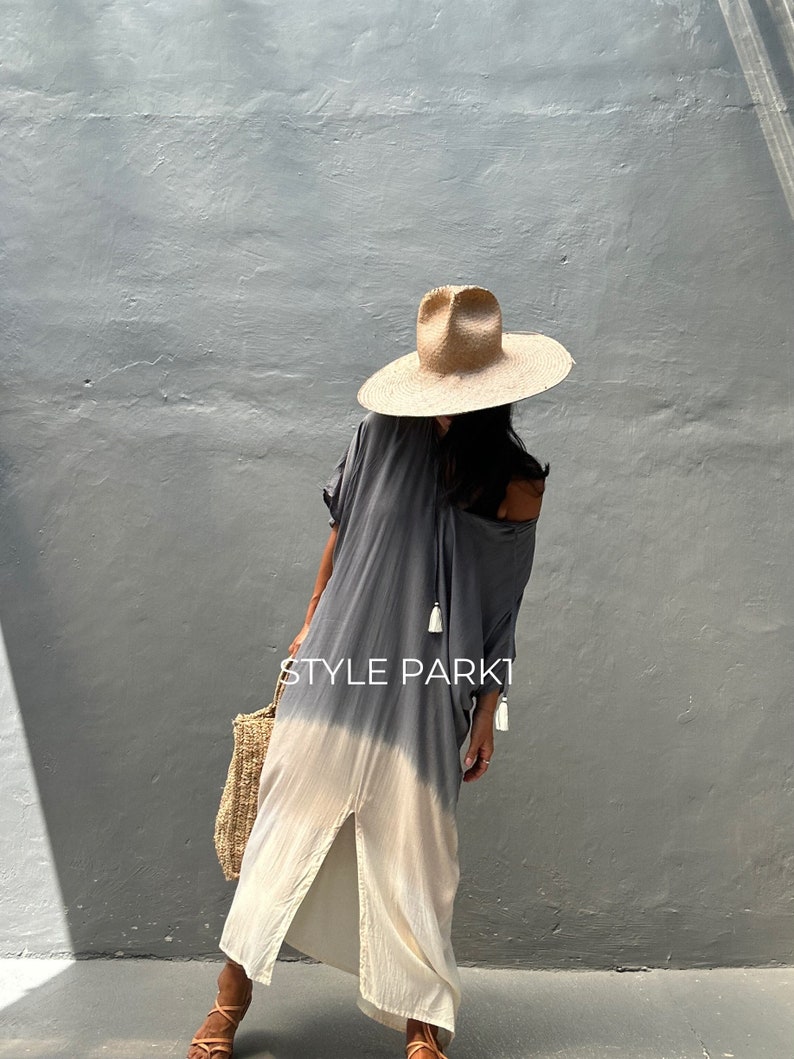 May include: A woman wearing a gray and white ombre maxi dress with a straw hat and a woven tote bag. The dress has a slit up the front and tassels on the neckline. The text "STYLE PARK1" is visible on the dress.