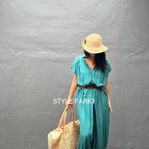 Peut inclure: Une combinaison bleu sarcelle avec une fermeture boutonn&eacute;e, des manches courtes et une taille cintr&eacute;e avec une ceinture marron. La tenue est accompagn&eacute;e d'un grand sac fourre-tout en paille, d'un chapeau de soleil et de sandales &agrave; lani&egrave;res. Le texte STYLE PARK1 est visible.