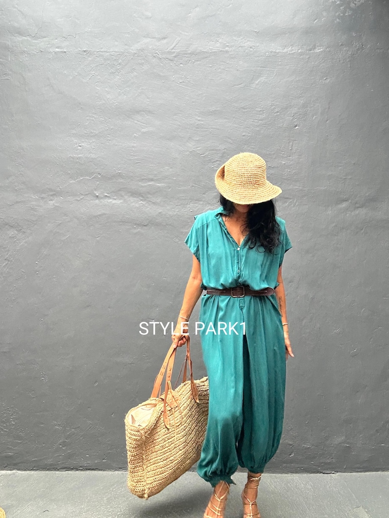 Peut inclure: Une combinaison bleu sarcelle avec une fermeture boutonn&eacute;e, des manches courtes et une taille cintr&eacute;e avec une ceinture marron. La tenue est accompagn&eacute;e d'un grand sac fourre-tout en paille, d'un chapeau de soleil et de sandales &agrave; lani&egrave;res. Le texte STYLE PARK1 est visible.
