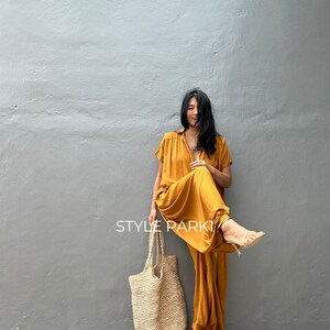 May include: A woman wearing a mustard yellow jumpsuit with a gathered hem and short sleeves. She is holding a large, natural fiber tote bag and smiling. The text "STYLE PARK" is visible in the image.