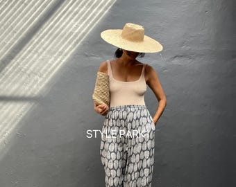 Könnte beinhalten: Eine Frau trägt ein beiges Tanktop und eine gemusterte Hose mit einem Strohhut und einer gewebten Tasche. Die Hose hat ein graues und weißes Blattmuster. Der Text "STYLE PARK" ist auf der Hose zu sehen.