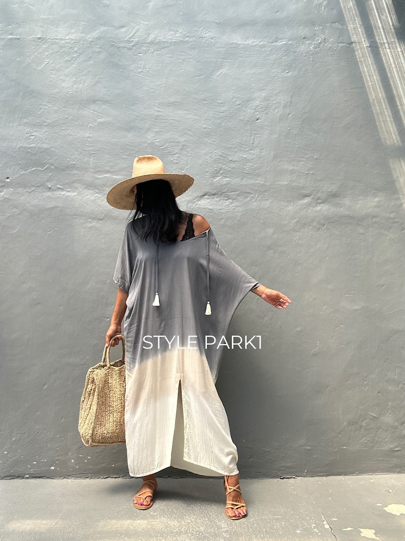 May include: A woman wearing a gray and white ombre maxi dress with a straw hat and a woven tote bag. The dress has a slit up the front and tassels on the neckline. The text "STYLE PARK1" is visible on the dress.