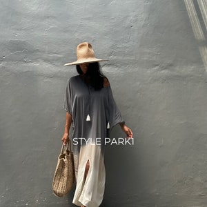 May include: A woman wearing a gray and white ombre maxi dress with a slit up the front. She is also wearing a wide-brimmed straw hat, a woven tote bag, and brown sandals. The text "STYLE PARKI" is visible on the dress.